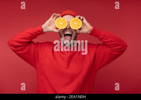 Photo of happy man with beard in red clothing covered eyes with orange fun tongue out, isolated over red background Stock Photo
