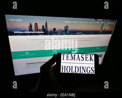 Person holding cellphone with business logo of Singaporean investment company Temasek Holdings on screen in front of website. Focus on phone display. Stock Photo