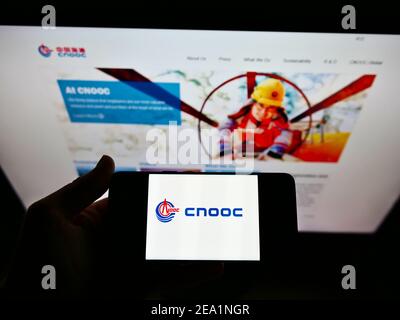 Person holding smartphone with logo of China National Offshore Oil Corporation (CNOOC) on screen in front of website. Focus on phone display. Stock Photo