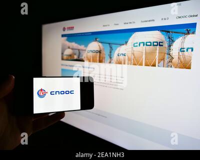 Person holding mobile phone with logo of China National Offshore Oil Corporation (CNOOC) on screen in front of web page. Focus on cellphone display. Stock Photo