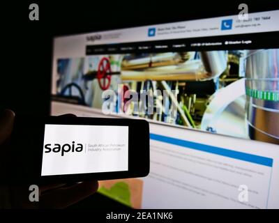 Person holding mobile phone with logo of South African Petroleum Industry Association (SAPIA) on screen in front of web page. Focus on phone display. Stock Photo