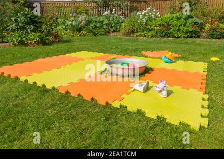 Color children's mat on the green grass in the country. Porous orange foam mat carpet. Children's shoes in the foreground Stock Photo