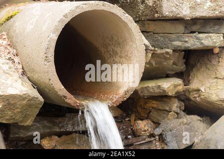 An old concrete drain pipe in a stone wall. A bit of water flows from the pipe. Focus is on the closest edge. Stock Photo