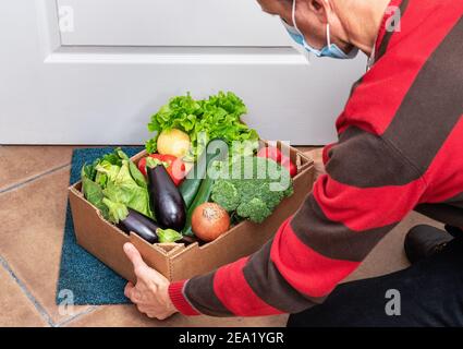 A delivery man puts a box of fresh vegetables on the floor in front of the door during quarantine and pandemic. Food supply concept. Stock Photo