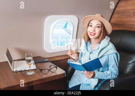 An Asian girl is flying on a plane in business class and drinking a glass of champagne, while working on a notebook online Stock Photo