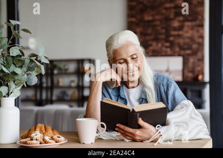 Smiling senior woman reading a book while sitting in the kitchen Stock Photo