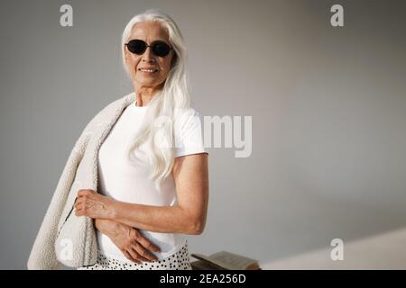 Joyful elderly white-haired woman in sunglasses smiling at camera isolated over grey background Stock Photo