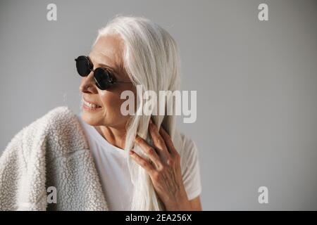 Joyful elderly white-haired woman in sunglasses smiling at camera isolated over grey background Stock Photo