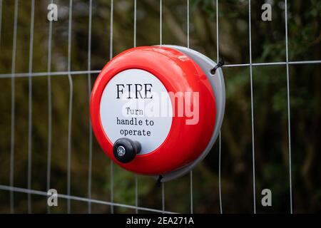Bright red metal fire alarm bell turn handle to operate alert manual steel ringing on construction building site attached to metal fence Stock Photo
