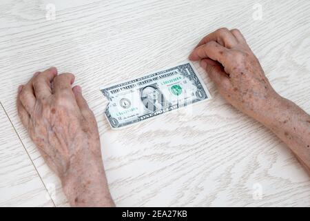 Hands of an elderly woman close-up lie on the table. Between them is 1 old ragged dollar. The concept of despair, poverty, lack of money for basic thi Stock Photo