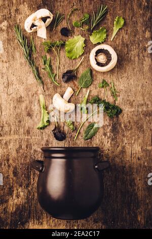 Layout of  fresh vegetables and mushrooms entering in the dark black pot against old wooden kitchen table background with nice, raw, texture. Nature s Stock Photo