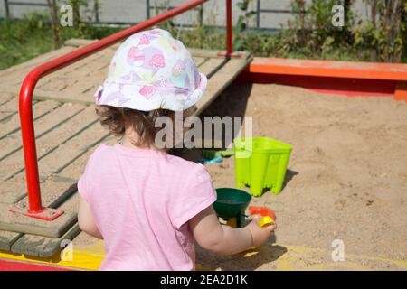 A little toddler girl in a panama hat on a bright sunny day plays in the sandbox . Concept summer in the city. Stock Photo