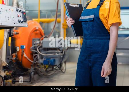 Concept of industrial production and factory. A female worker in uniform with a folder in her hand. In the background, boiler equipment. Stock Photo