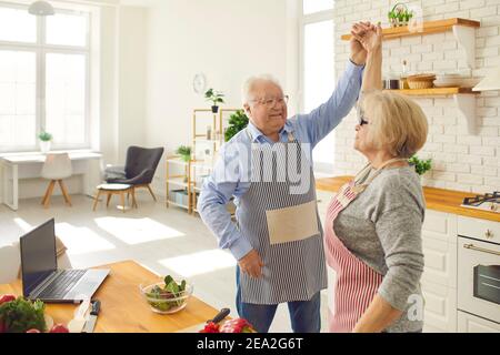 Happy senior woman dancing with mature loving husband in the kitchen while cooking dinner.