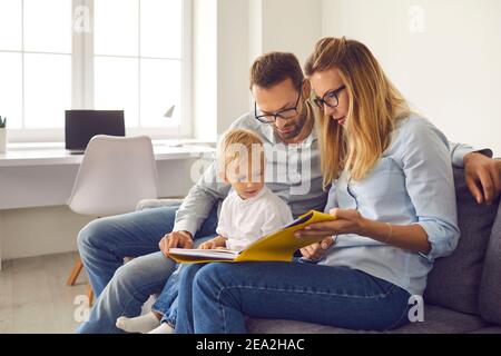Mom, dad and son read a book of stories together sitting on the couch in a bright room at home.