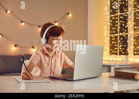 Red haired boy in headphones sitting, watching lesson or course online on laptop and making notes Stock Photo