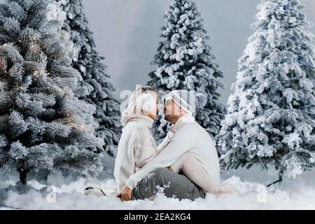 Man kiss and hug his woman and snow falls. New year love story. People weared wearing fur headphones, hats, white sweaters. Stock Photo