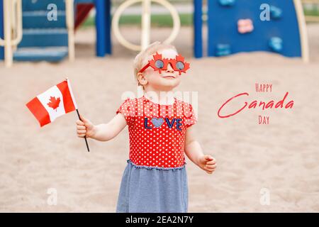 Happy Canada Day. Holiday card with text. Funny baby toddler girl holding waving Canadian flag. Kid child wearing funny maple leaves sunglasses Stock Photo