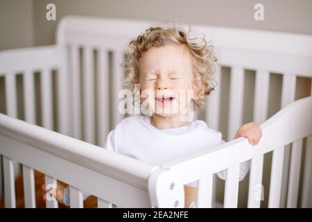 Cute adorable baby boy crying in crib at kids nursery room at home. Funny baby boy with curly blond hair screaming at his bed. Terrible two toddler ch Stock Photo