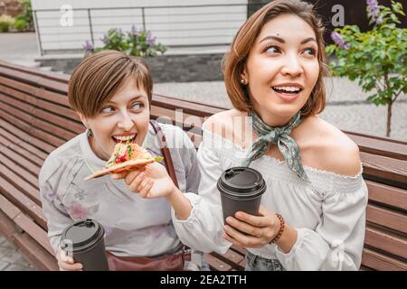 Girl friends share a takeaway pizza. They eat delicious fast food and drink coffee and socialize in a cozy city square Stock Photo