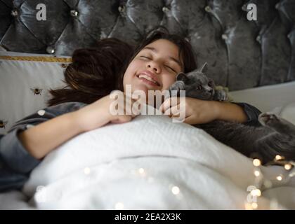 Smiling girls in grey pyjamas lying and slipping on the bed and hugging a cat in the morning Stock Photo