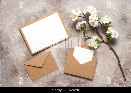 Blank wedding card mockup in brown envelopes and cherry blossom branch (artificial). Beige grunge background. Spring stationery still life. Top view,