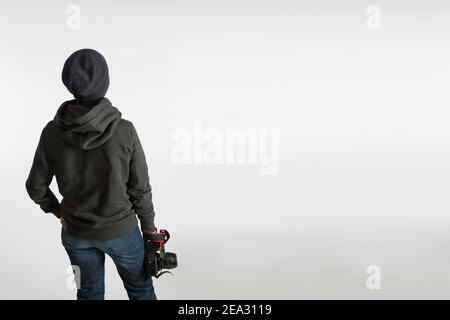 A teenager with a SLR camera on his back stands in a hoodie and a hat on a blue background. Photography training concept Stock Photo
