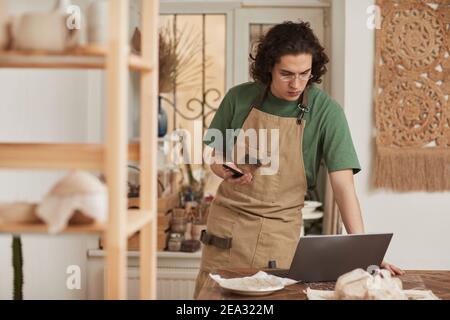 Waist up portrait of young artisan using laptop in crafting workshop, small business concept, copy space Stock Photo