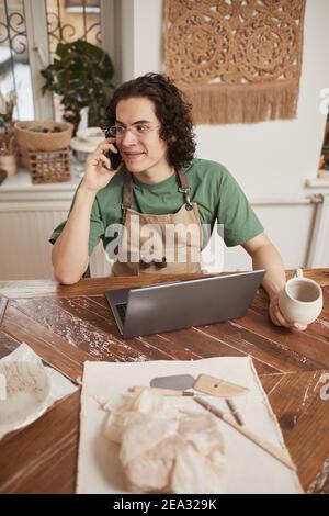 Vertical portrait of young man speaking by smartphone and using laptop in workshop while managing small business, copy space Stock Photo