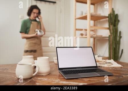 Still life of laptop with blank white screen on table in workshop, with blurred male artisan speaking by phone in background, small business and entre Stock Photo