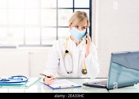 Shot of female doctor wearing face mask for prevention while sitting at desk and making a call in the doctor’s office. Stock Photo
