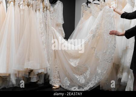 Bride touches luxurious white cream wedding dress on hangers. Dresses made of silk chiffon, tulle and lace. Luxury pearls and crystals pendants on the Stock Photo