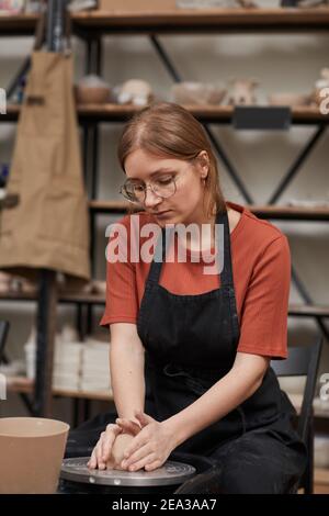 Vertical warm-toned portrait of young woman shaping clay on pottery wheel while enjoying hobby in workshop Stock Photo
