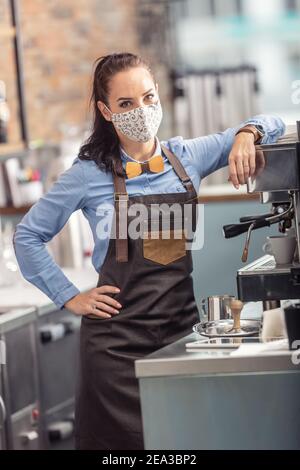 Fashionable waitress wears protective face mask while preparing an espresso in a coffee house. Stock Photo