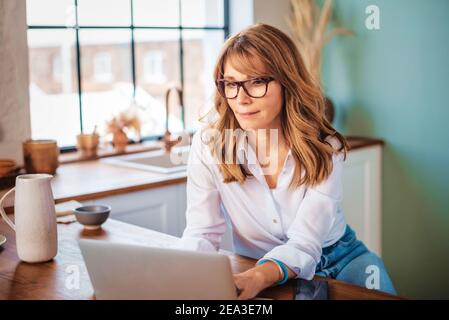 An attractive middle aged woman using her laptop while relaxing at home in her kitchen. Stock Photo