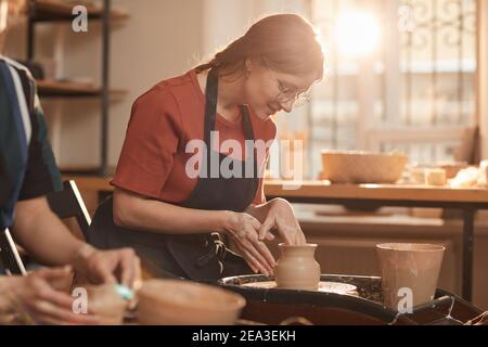 Warm toned portrait of young woman shaping clay on pottery wheel in sunlit workshop and enjoying arts and crafts, copy space Stock Photo