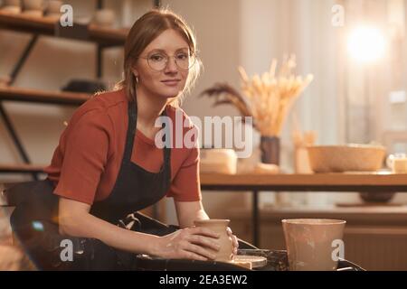 Warm toned portrait of young woman shaping clay on pottery wheel while making ceramics in sunlit workshop, copy space Stock Photo