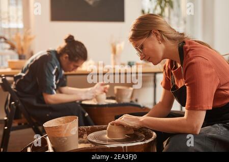 Side view portrait of young woman shaping clay on pottery wheel while making ceramics in workshop lit by sunlight, copy space Stock Photo