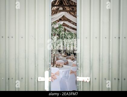 Sneaking a look at wedding reception party laid out behind rustic doors in traditional old converted farm barn with blossom trees white tables Stock Photo