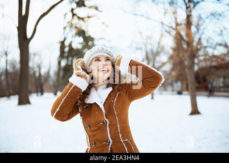 happy stylish female with mittens in a knitted hat and sheepskin coat outside in the city park in winter. Stock Photo