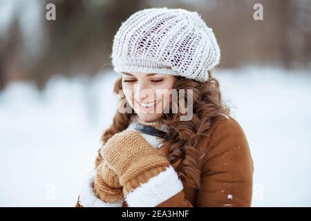 happy stylish middle aged woman with mittens and cup of hot cocoa in a knitted hat and sheepskin coat outdoors in the city park in winter. Stock Photo