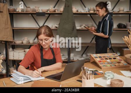 Portrait of female small business owner taking notes in planner while working at wooden table with laptop in pottery workshop, copy space Stock Photo