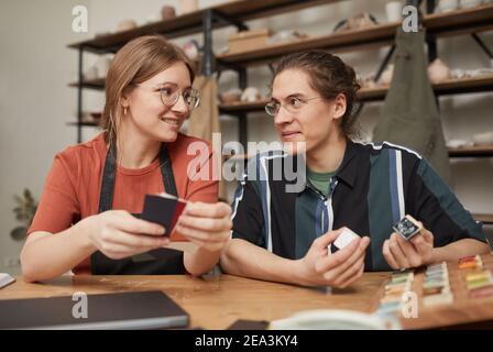 Portrait of two young artists choosing color samples and smiling while sitting at wooden table in pottery workshop Stock Photo