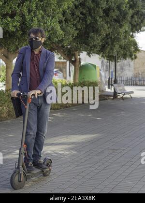 A shallow focus of a young businessman wearing a facemask and going to work with a small scooter Stock Photo