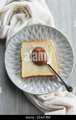 Plate with spoonful of hazelnut praline paste on slice of bread,white towel and grey table as background Stock Photo
