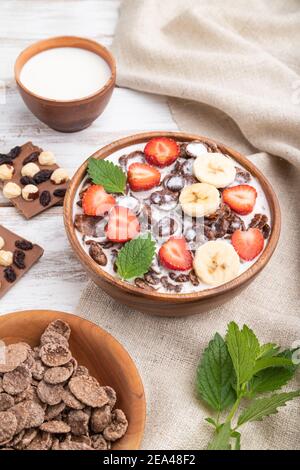 Chocolate cornflakes with milk and strawberry in wooden bowl on white wooden background and linen textile. Side view, close up. Stock Photo
