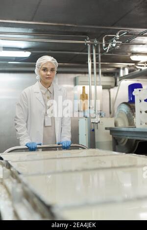 Vertical portrait of young female worker pushing cart with raw cheese in workshop at food factory and looking at camera, copy space Stock Photo