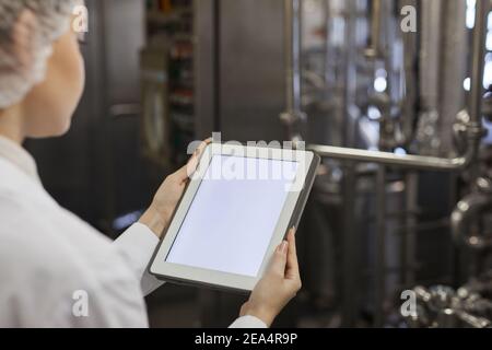Close up of young female worker holding digital tablet with blank white screen against factory equipment in background, copy space Stock Photo
