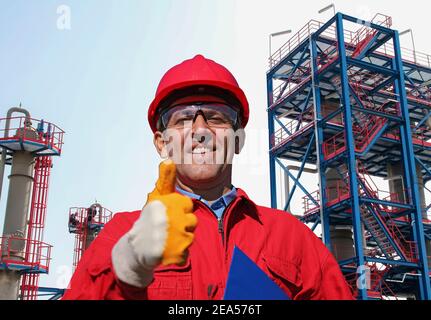 Happy Oil Worker in Red Hardhat Giving Thumb Up in front of Oil and Gas Refinery. Portrait Of Smiling Man in Protective Workwear. Stock Photo