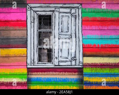 Vintage window, painted with white and black colors, in the colorful peeled wall wooden old house. Fragment of ancient, multicolored, textured wall. Stock Photo